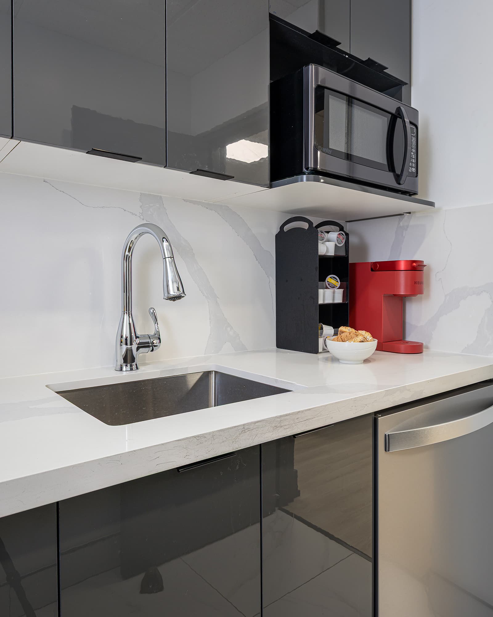 Modern kitchen with glossy grey cabinets, white quartz countertop, stainless steel sink, and red coffee maker beside built-in microwave.